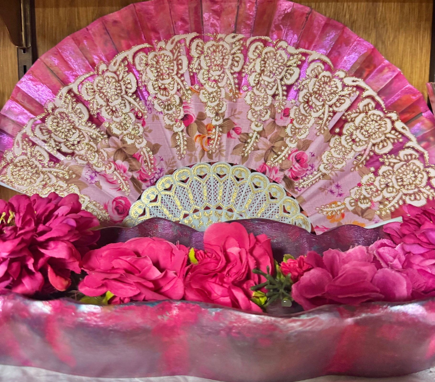 A pink and gold lace fan resting above fuchsia roses on an altar