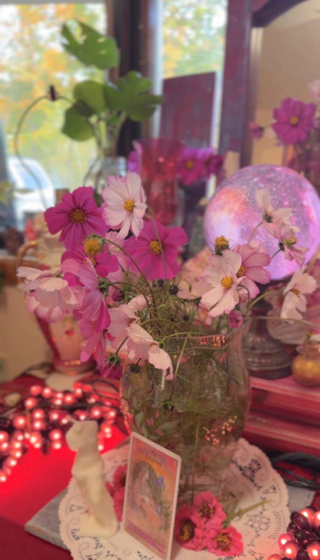 A white Venus figure among pink cosmos before a glowing moon orb
