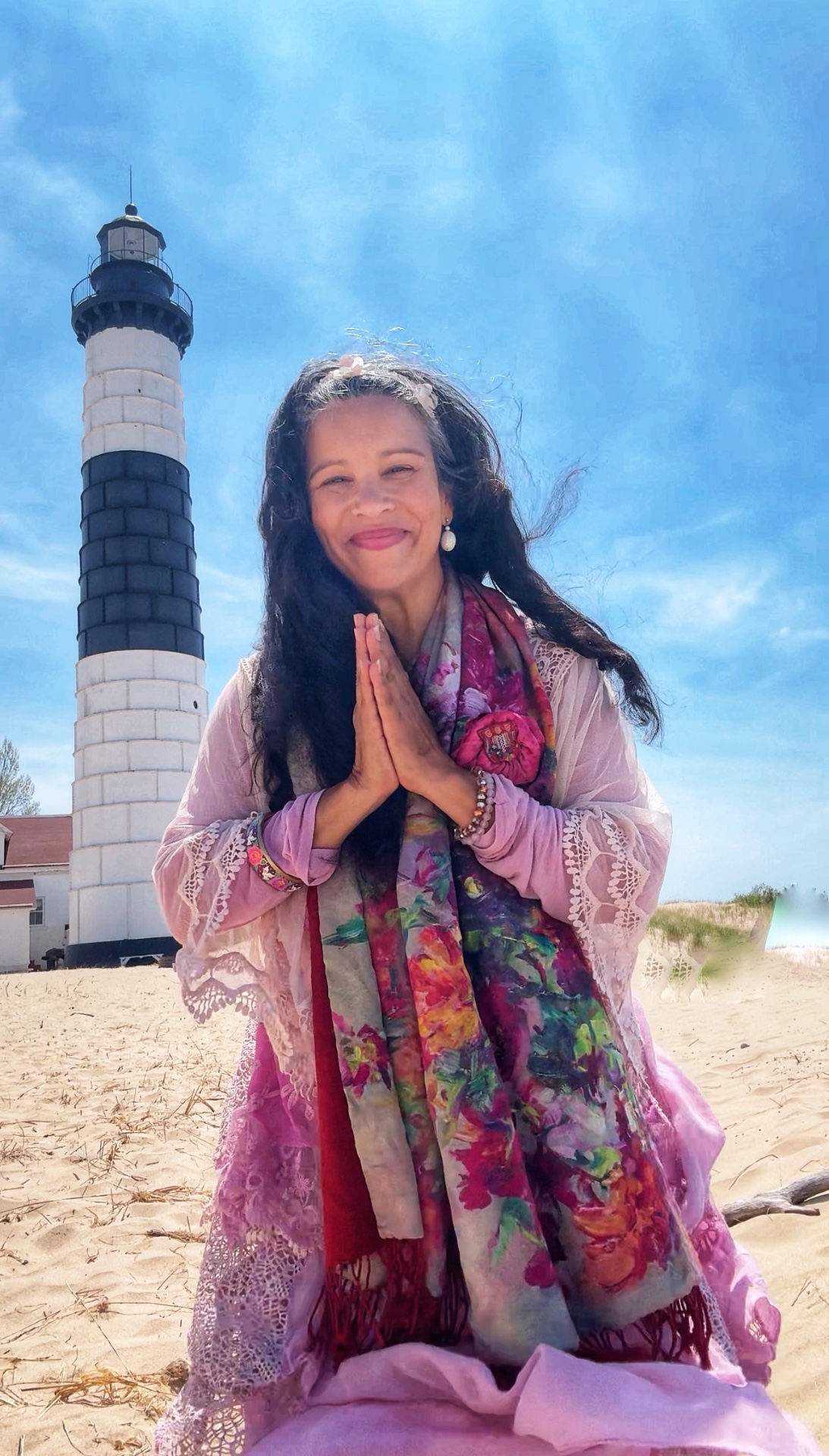 Reverent Mariela in pink silks and lace, hands in prayer on the beach beside a lighthouse