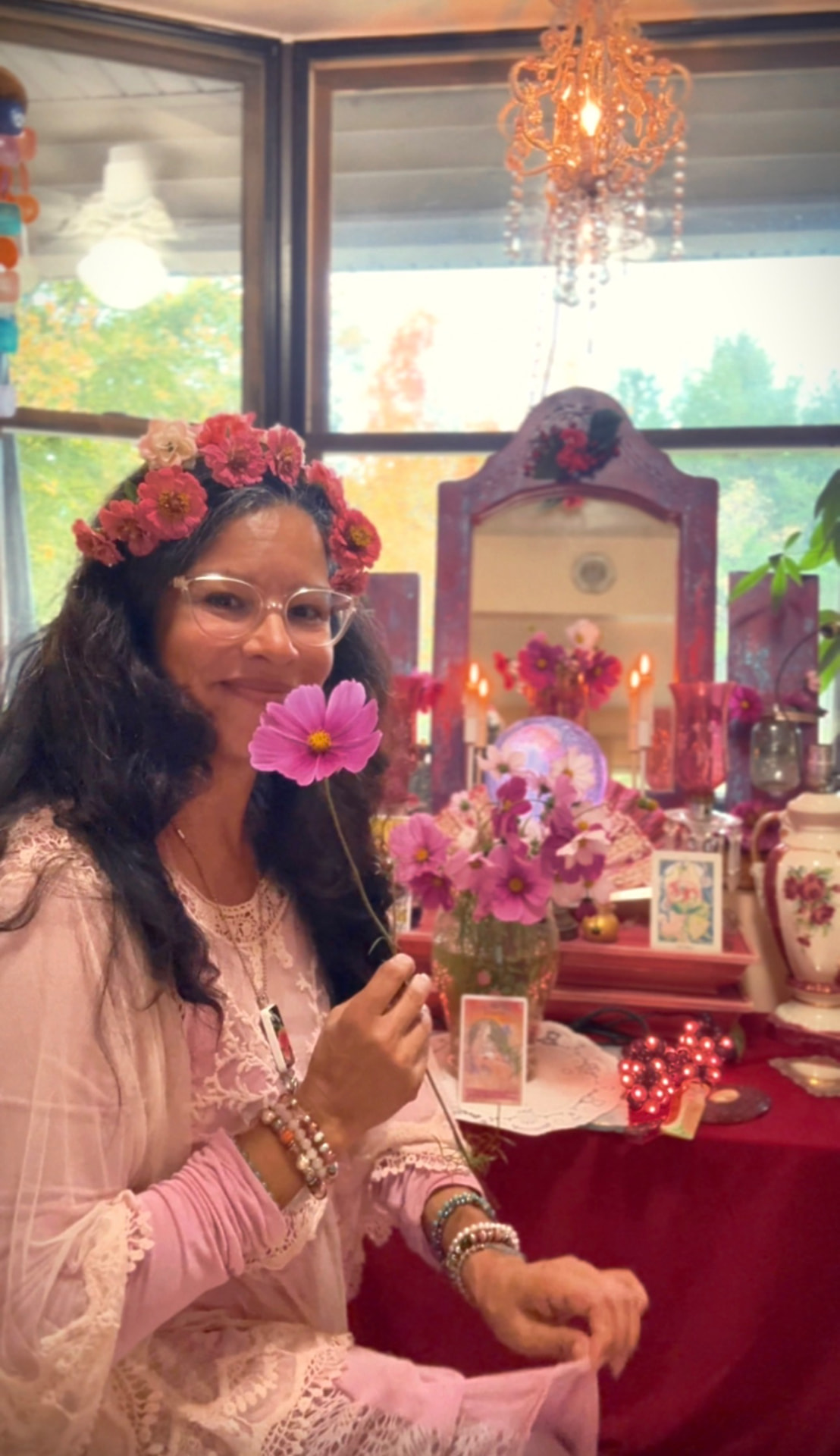 Reverent Mariela in a crown of zinnias, holding a pink cosmos flower before her rose altar