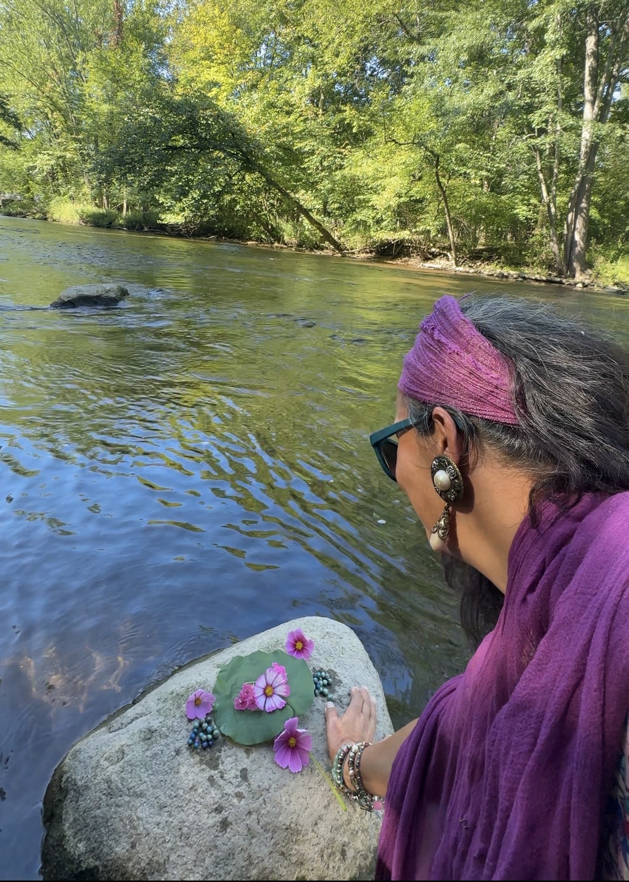 Mariela at the river's edge, her hand resting on a stone offering of cosmos flowers on a lily leaf