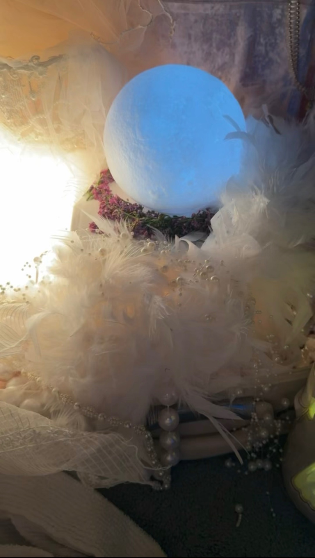 A soft blue moon nested in white feathers, lilacs, and pearls