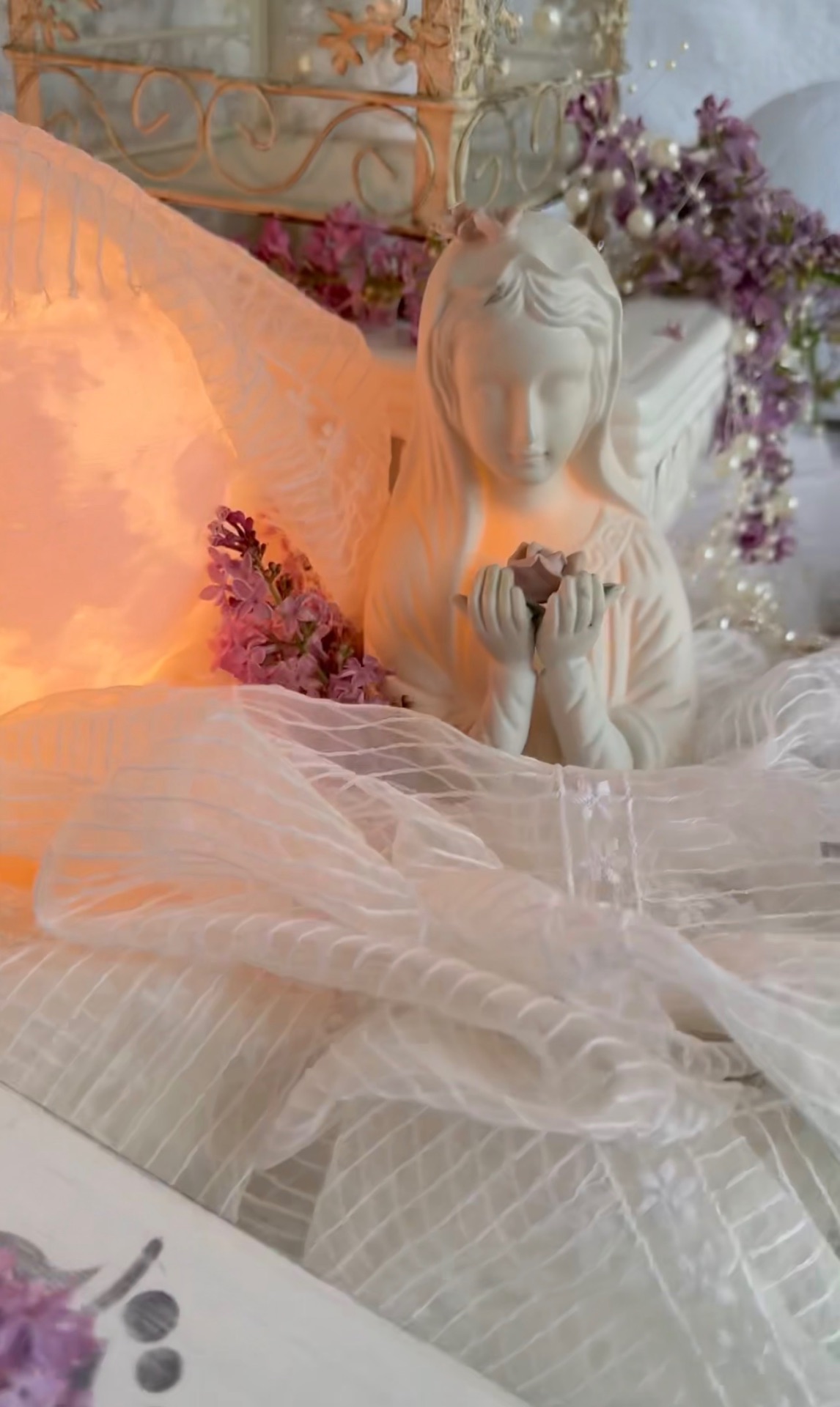 A white ceramic figure of a woman holding a flower bud, beside a glowing lamp, lilacs, pearls and an antique wire cage