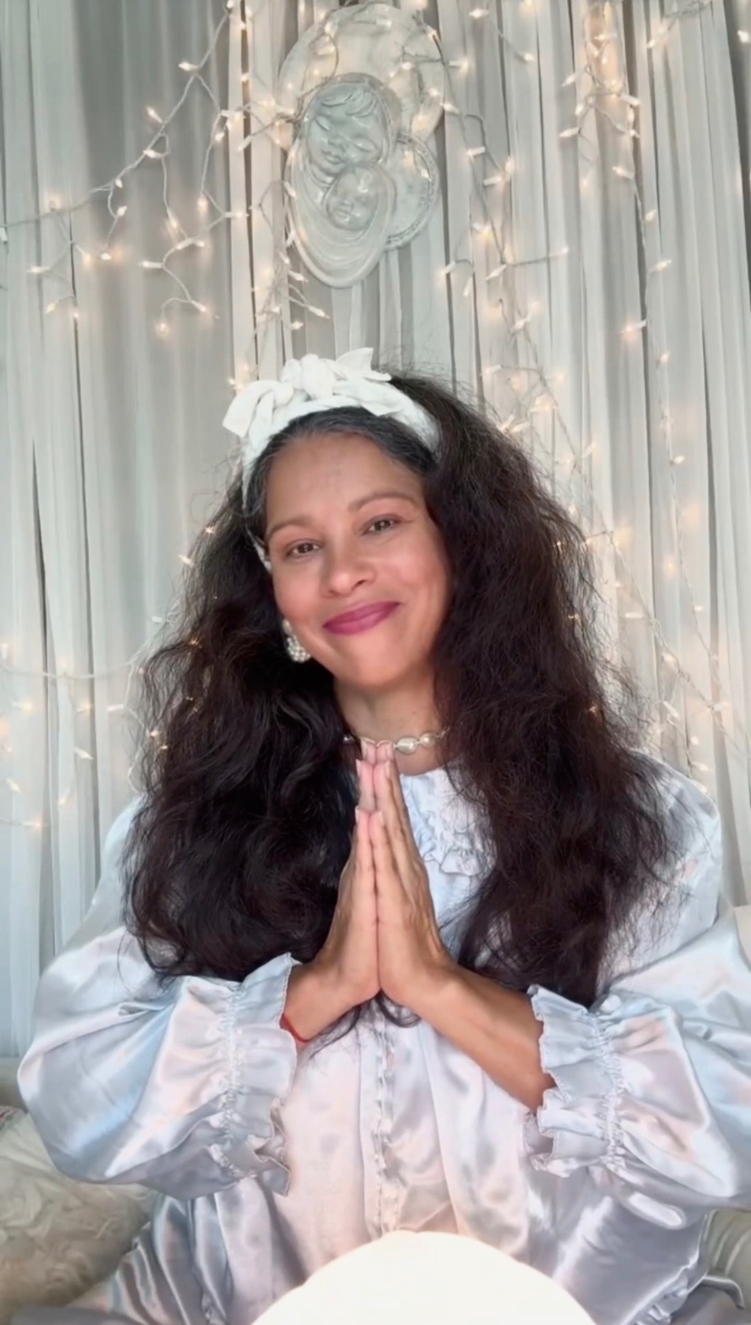 Mariela in white satin, hands joined in reverence before softly lit curtains