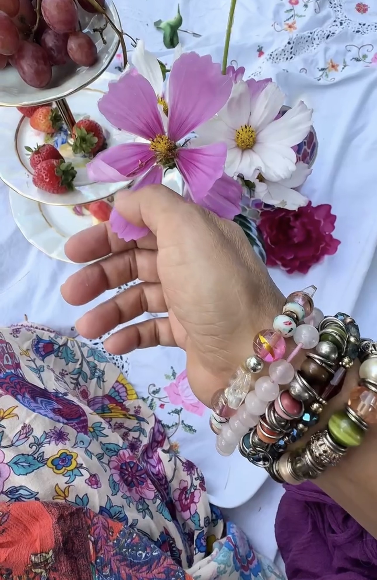 A hand wearing beaded bracelets reaching toward cosmos flowers on a lace tea table