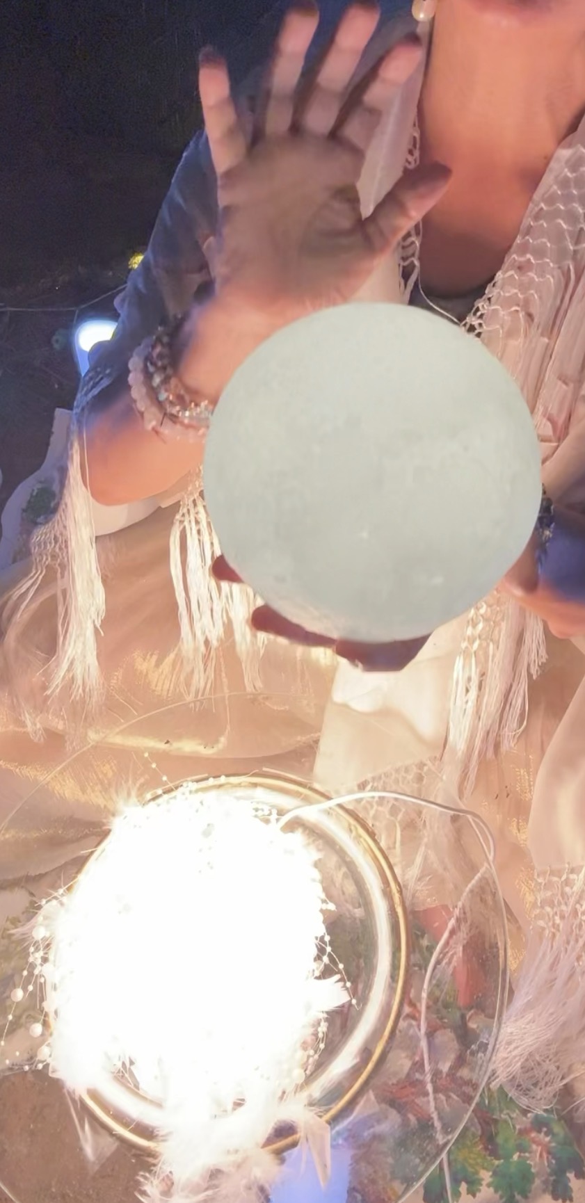 Mariela cradling a glowing moon orb in her hands above a bowl of feathers, pearls and light