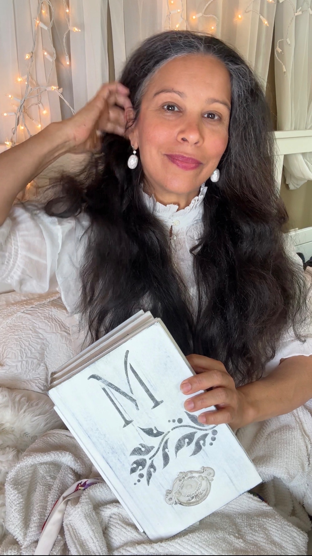 Mariela in white lace on her bed beneath fairy lights, holding a white book marked with an M, a Mother Myth