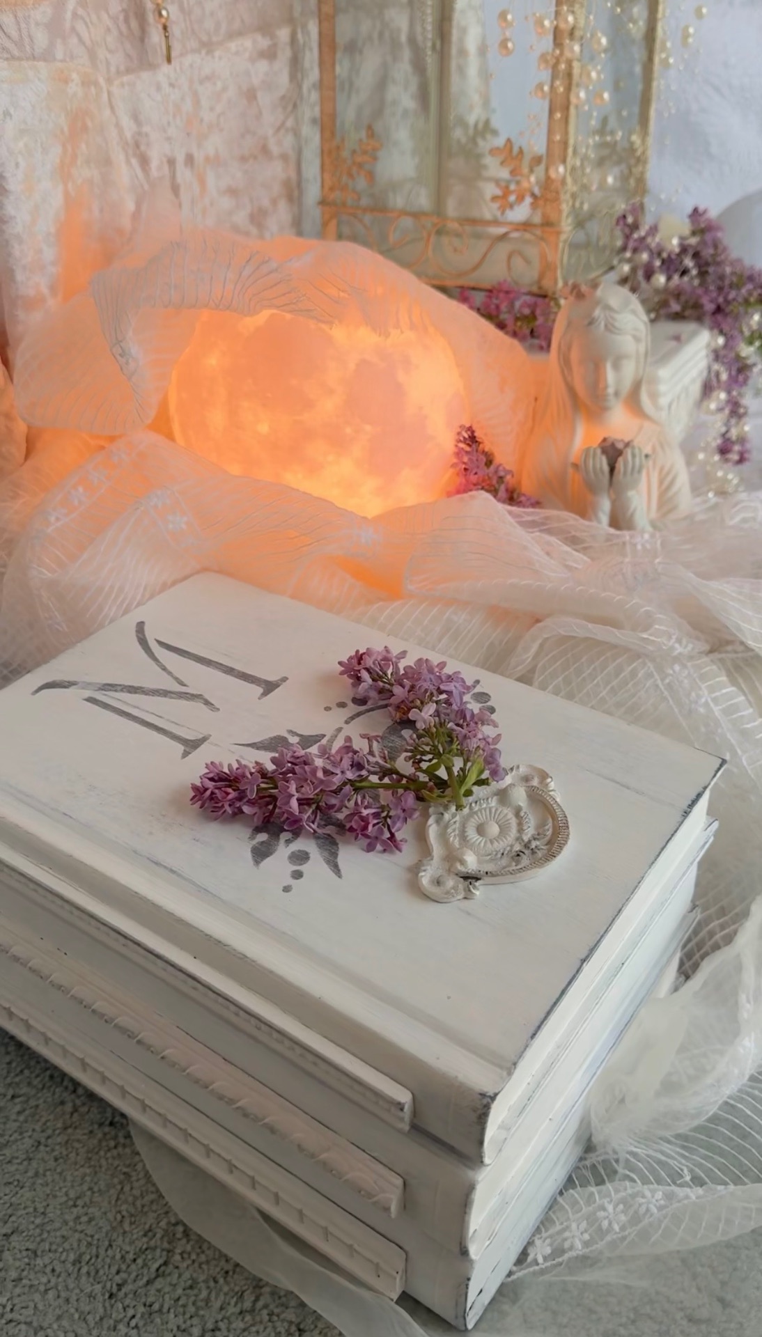 A glowing moon resting in tulle behind white books adorned with lilacs