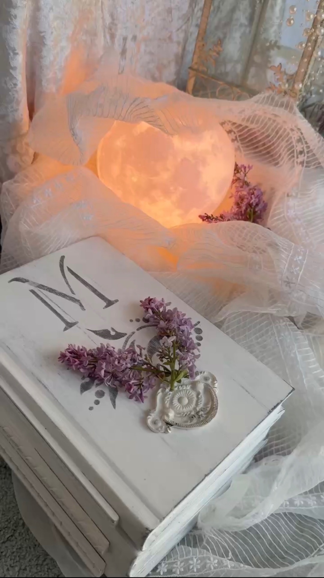 A white book box monogrammed with M and lilac blossoms before a glowing moon lamp nestled in lace