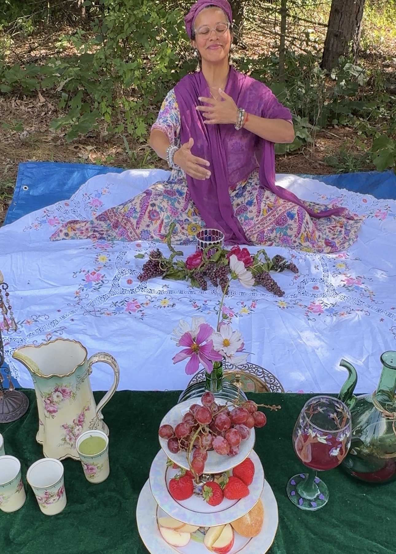 Mariela seated at a forest table set with tea, fruit, and flowers
